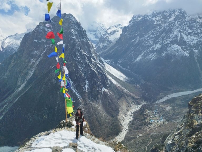 A trekker standing near prayer flags with a panoramic backdrop of the Himalayan mountain range and a distant village in the valley.