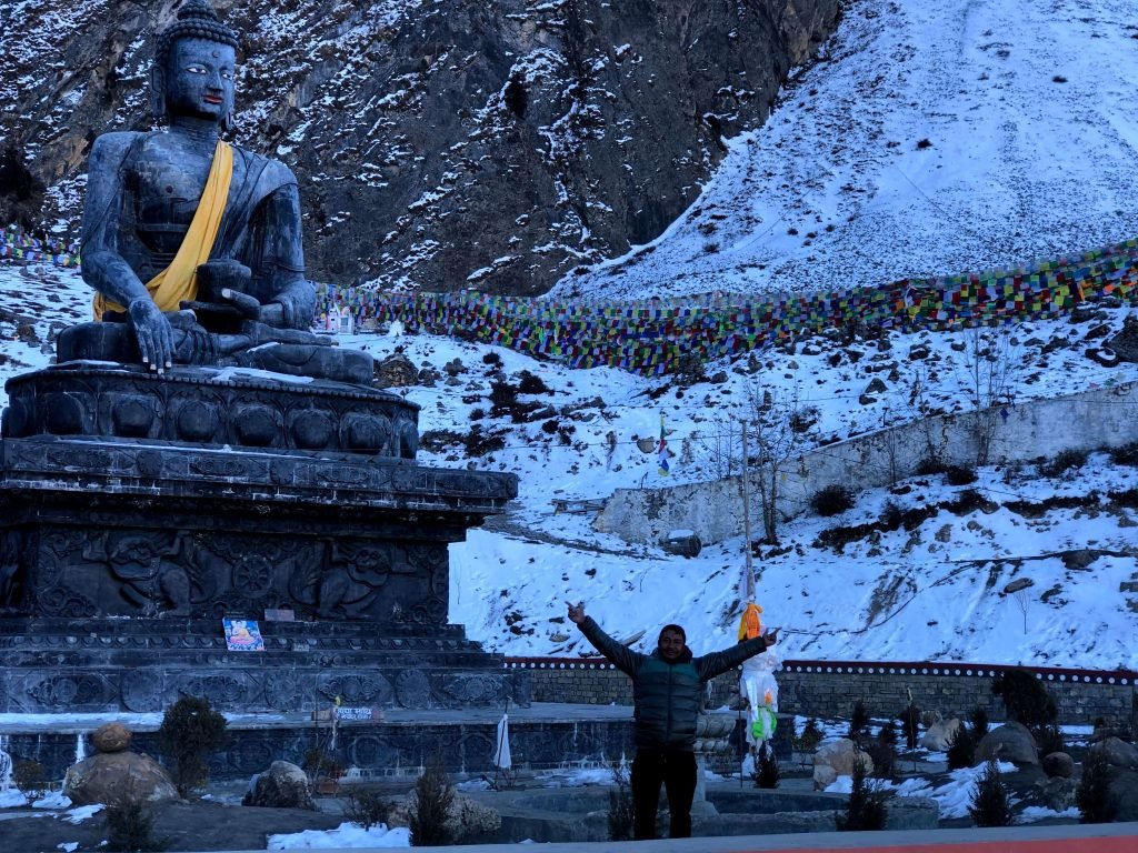 A majestic Buddha statue draped in a yellow cloth overlooks the snowy landscape near Muktinath Temple. A joyful traveler stands below, arms outstretched, amidst prayer flags and rugged mountains.