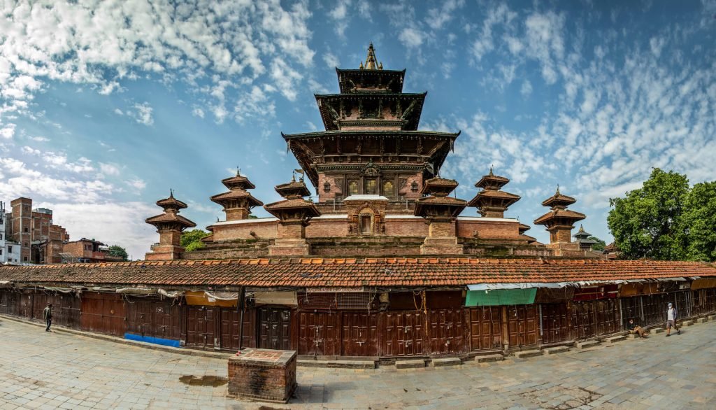 A panoramic view of an ancient multi-tiered temple with smaller temples surrounding it, set against a blue sky with scattered clouds. In the foreground, a series of wooden stalls and a few individuals can be seen walking on the paved ground. Best Time to Visit Nepal
