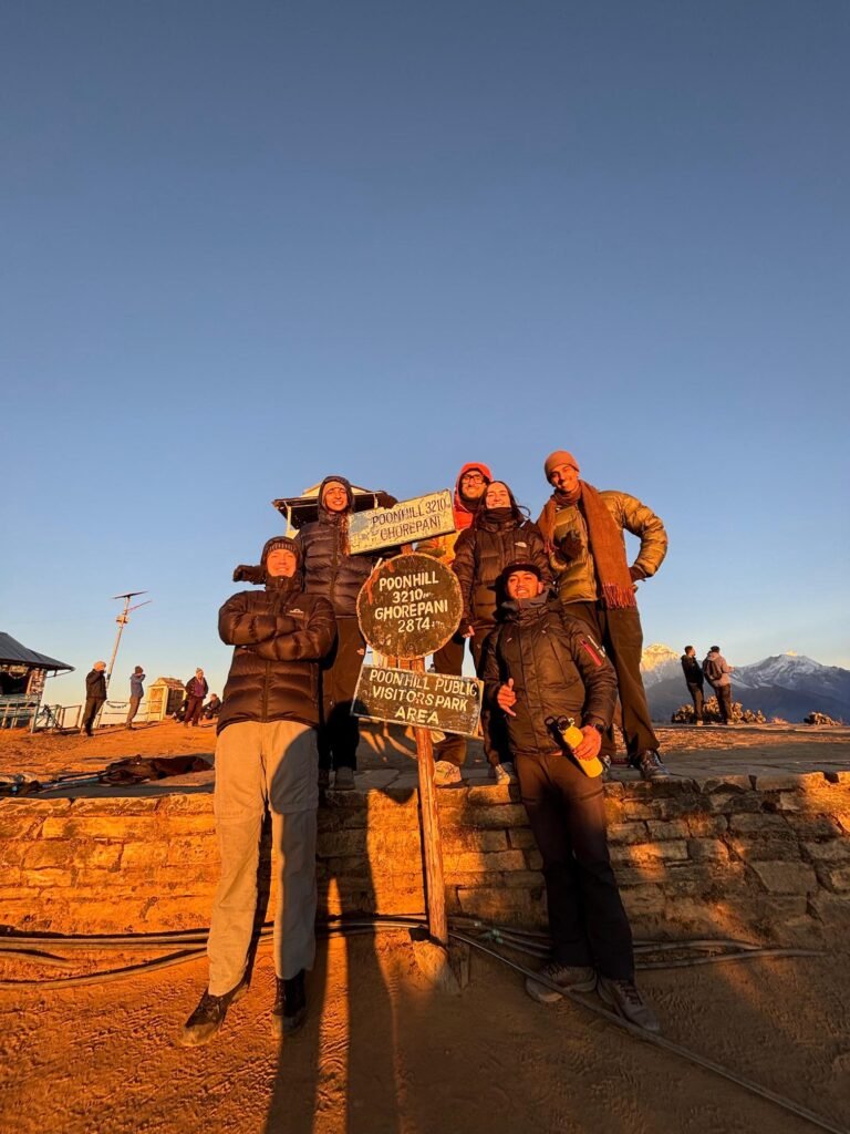 Trekkers at sunset near Ghorepani Poonhill signpost with mountain views.