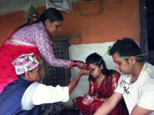 A candid photograph capturing a traditional Nepali blessing ceremony indoors. A woman in a pink and red patterned top and red sari pours something from a small metal vessel onto the hand of an elderly man, who in turn applies a red mark (tika) to the forehead of a seated young woman in a red sari. A young man with green grass sprigs in his hair sits next to her, also receiving a blessing. The background shows simple, earthy-toned walls and traditional indoor settings.