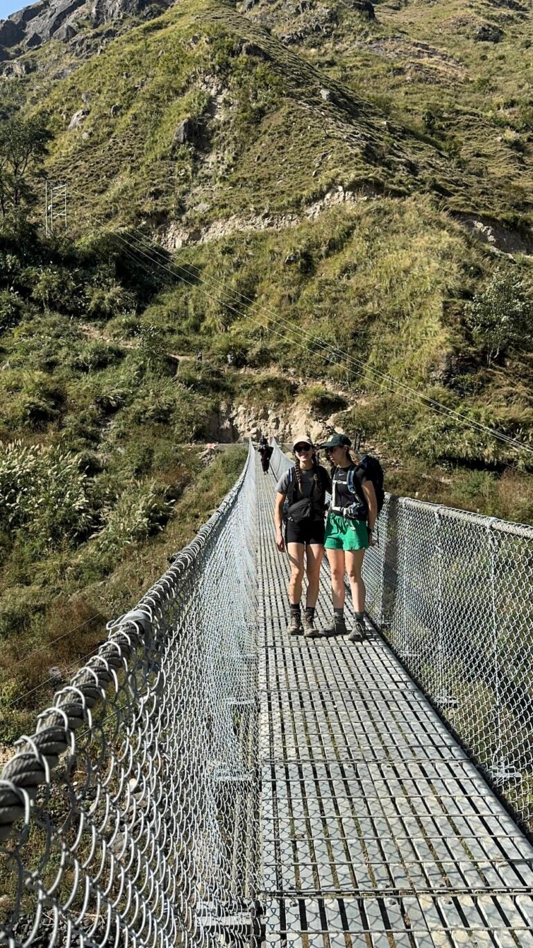 Two hikers crossing a steel suspension bridge between green hills on the Manaslu Circuit Trek near Filim, Nepal