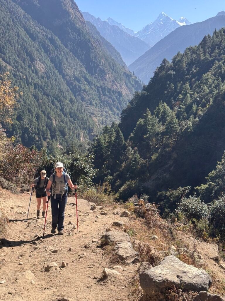 Hiker ascending a rocky mountain trail to Lho on Nepal's Manaslu Circuit Trek with pine forests and Himalayan peaks.